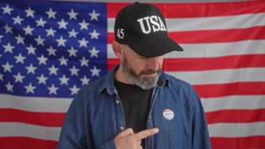 Amerikan bayrağının önündeki şapkalı, gülümseyen sakallı adam, oy verdikten sonra işaret ediyor ve işaret ediyor.