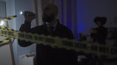 Sarı bant ve el feneriyle loş aydınlatılmış bir suç mahallini araştıran bir adam ve kadın.