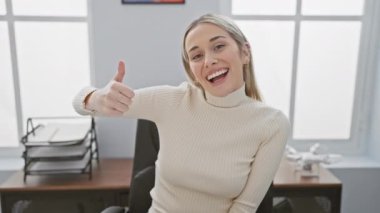 Ofis masasında oturan neşeli, genç, sarışın bir kadın. Dostane bir gülümseme ve olumlu bir tavırla ışıldıyor. Havalı bir onay ve başarı işareti gösteriyor.