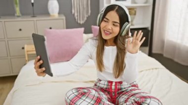 Pijamalı genç bir İspanyol kadın rahat bir yatak odasında, yatak odasında, kulaklık takmış bir tabletle selfie çekiyor..