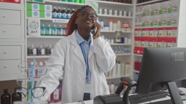Genç bir Afrikalı Amerikalı kadın eczacı eczanede telefonla konuşuyor. İlaç raflarıyla dolu..