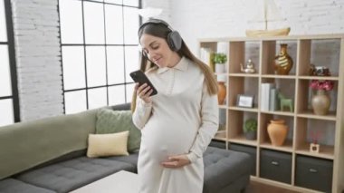 Kulaklıklı hamile bir kadın rahat bir oturma odasında akıllı telefon kullanıyor, modern annelik ve ev içi rahatlığı somutlaştırıyor..