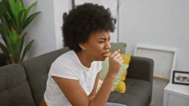 Rahat bir oturma odasında gri bir kanepede otururken içeride öksüren Afrikalı Amerikalı kadın..