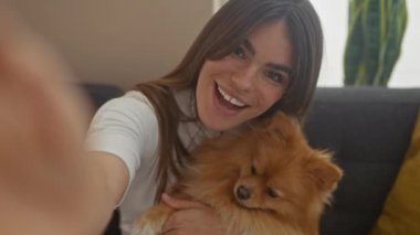 Sıcak bir odada gülümseyen bir kadın sevimli Pomeranian köpeğiyle selfie çekiyor.