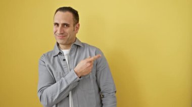 Tişörtlü neşeli, genç, İspanyol bir adam, doğal olarak gülümsüyor ve elini ve parmağını havaya kaldırıyor. Yüzünde mutlu bir ifade, yalıtılmış sarı bir arkaplanda duruyor.