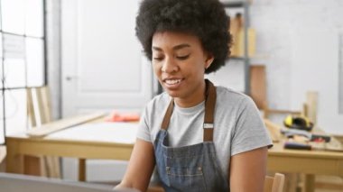 Marangozluk aletleri arka planda dinlenirken, bir atölyede dizüstü bilgisayar kullanan odaklı siyah bir kadın..