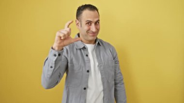 Tişört giyerken küçük boy işaret veren, sarı izole edilmiş arka planda duran mutlu ve çekici parmaklarını çimdikleyen kendine güvenen İspanyol genç adam. Ölçüm konsepti.