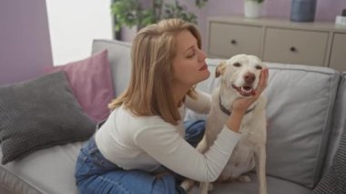 Sarışın bir kadın evdeki kanepede beyaz köpeğini sevgiyle öpüyor, sevgisini ve dostluğunu gösteriyor..