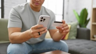 Asyalı bir adam modern bir oturma odasında akıllı telefonunu kullanırken kredi kartını şüpheyle inceliyor..