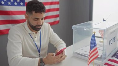 Sakallı genç bir İspanyol, oy sandığı ve bayrağı olan bir Amerikan oy kullanma merkezinde telefonunu inceliyor..