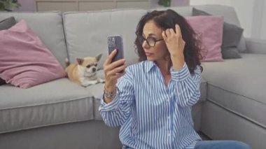 Akıllı telefonu ayna olarak kullanan olgun bir kadın ve sıcak oturma odasında Chihuahua köpeği.