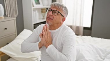 Umutsuz dua, orta yaşlı, pijamalı gri saçlı adam, umuda tutunuyor, yatak odasında el ele yalvarıyor.