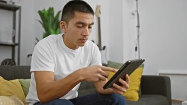 İspanyol adam modern bir oturma odasındaki koltukta tablet kullanıyor. Bağlantı ve günlük yaşam tarzını aktarıyor..