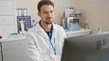 Laboratuvarda gözlüklü, dizüstü bilgisayarlı ve beyaz önlüklü genç bir İspanyol profesyonellik saçıyor ve tıbbi bir araştırma ayarına odaklanıyor..