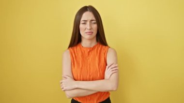 Şüpheli, kolsuz tişörtlü negatif güzellik, çapraz kollu duruş, kapalı alanda sarı arka planda gergin ve onaylanmayan ifade