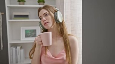 Yatak odasında, elinde kahve fincanı tutarken kulaklıkla müzik dinleyen rahat bir genç kadın..