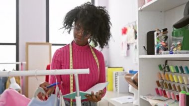 Terzi dükkanındaki Afrikalı Amerikalı kadın arka planda kumaş ve dikiş aksesuarlarıyla notlar alıyor..