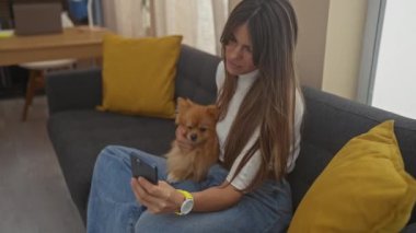 Uzun saçlı gülümseyen genç bir kadın, gri bir kanepede oturuyor. Pomeranian köpeğine sarılıyor ve akıllı telefonu içeride tutuyor..
