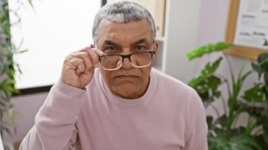 Gözlüklü, neşeli, gri saçlı bir adam ofis ortamında neşeli bir ifadeyle poz veriyor..