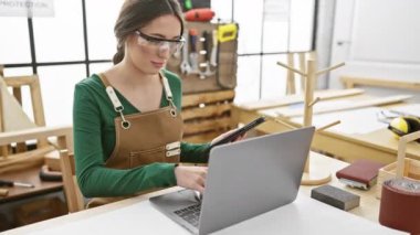 Önlüklü kadın ahşap atölyesinde dizüstü bilgisayar ve aletlerle tablet kullanıyor.