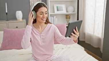 Pijamalı ve kulaklıklı bir kadın yatağında tablet kullanıyor, rahat, modern yatak odası dekorunu tasvir ediyor..