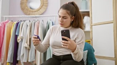 Soyunma odasında genç bir kadın makyajını aynası ve akıllı telefonuyla kontrol ediyor. Etrafı renkli kıyafetlerle çevrili..