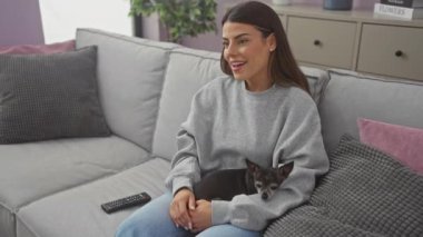 Gülümseyen bir kadın evcil hayvanıyla kanepede uzanmış, rahat bir oturma odasında, televizyon izlerken sevinçlerini dile getiriyor..