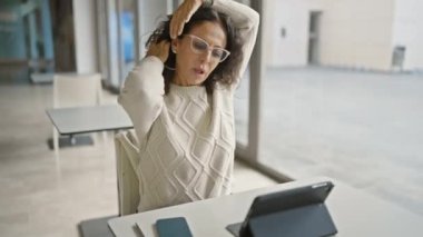 Gözlüklü ve süveterli olgun bir İspanyol kadın elinde tablet ve akıllı telefonuyla bir ofis masasında dinleniyor..