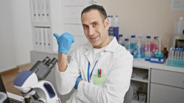 Bilim adamı üniforması içinde neşeli, genç İspanyol bir adam laboratuarda çalışmaktan keyif alıyor, başparmakları havada ve yüzünde kocaman bir gülümsemeyle kenarı işaret ediyor.!