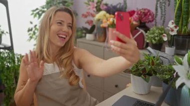 Neşeli sarışın bir kadın çiçek ve buketlerle dolu canlı bir çiçekçide selfie çekiyor. Küçük işletme sahibinin gururunu okşuyor..