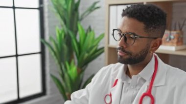 Klinikte sakallı, gözlüklü ve stetoskop takmış, iş yerinde gülümseyen kendinden emin, Afro-Amerikan bir erkek doktor..