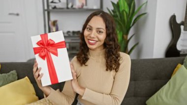 Neşeli ve kendinden emin genç Afrikalı Amerikalı kadın doğum günü hediyesini sıkıca tutuyor, evinin sıcacık iç kısmında dişlek gülümsemesini sergiliyor..