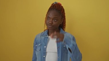 Kendinden emin, çekici Afrikalı Amerikalı kadın gülümsüyor ve şık bir kot gömlekle poz veriyor, saçlarına dokunuyor, izole edilmiş sarı arka planda duruyor.