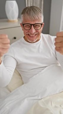 Orta yaşlı, gri saçlı bir adam mutlu, başarılı bir baş parmak işareti yapıyor, neşeli bir gülümsemeyle parlıyor. Pijama giyerse, onun jesti kazananın neşesidir.