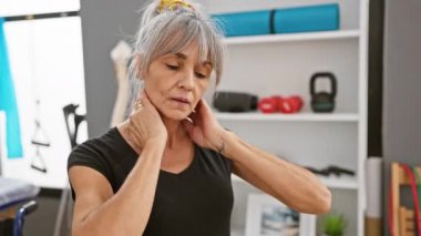 Gri saçlı olgun bir kadın rehabilitasyon kliniğinde boyun ağrısı çekiyor. Rahatsızlık ve sağlık hizmetlerini tasvir ediyor..