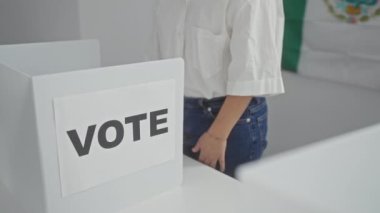 Orta yaşlı bir kadın, Meksika bayrağının önünde, kapalı alanda oy veren bir oy kabininin yanında kollarını kavuşturarak duruyor..