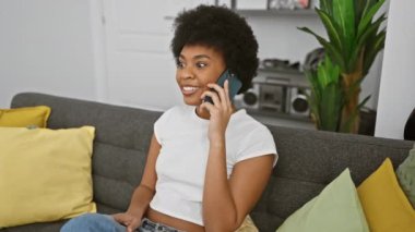 Hoş bir oturma odasında renkli yastıklarla akıllı bir telefonla gülen neşeli Afrikalı kadın..