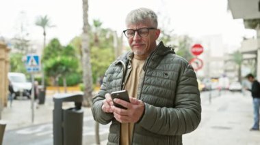 Gözlüklü ve gri saçlı olgun bir adam şehir sokaklarında akıllı telefon kullanır. Şehir bağlantısını ve modern yaşam tarzını somutlaştırır..