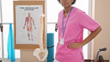 Afro-Amerikan kadın sağlık uzmanı anatomi çizelgesi ve iskelet modeli ile klinikte duruyor..
