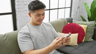 Modern evdeki koltukta akıllı telefon kullanan gülümseyen Asyalı adam..
