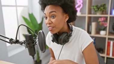 Modern bir radyo stüdyosunda mikrofona konuşan kıvırcık saçlı, kulaklık takan neşeli bir kadın..