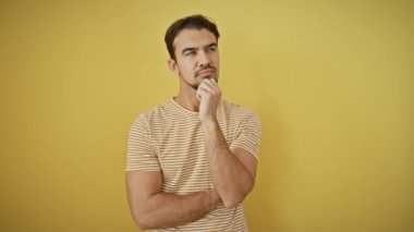 Çizgili tişört giyen düşünceli genç İspanyol adam, sarı izole edilmiş bir arka plana karşı duruyor. Ciddi yüzü, eli çenesinde, derin düşüncelere dalmış, kafa karıştırıcı bir fikri var..