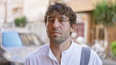 Çekici, sakallı ve yeşil gözlü İspanyol bir adam güneşli bir şehir sokağında dikiliyor..