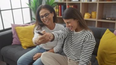 İki kadın rahat bir salonda, rahat bir koltukta uzaktan kumanda için gülüyor ve şakalaşıyor..