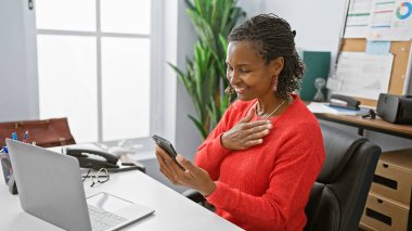 Ofiste akıllı telefon kullanırken gülümseyen Afrikalı Amerikalı kadın, bir iş ortamında üretkenlik ve teknolojiyi ön plana çıkarıyor.