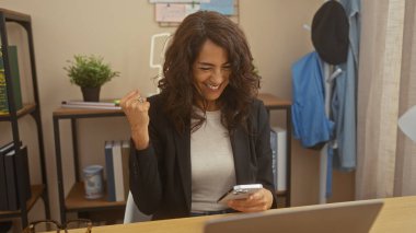 Gülümseyen orta yaşlı bir kadın ev ofisindeki başarısını akıllı telefon ve dizüstü bilgisayarla kutluyor..