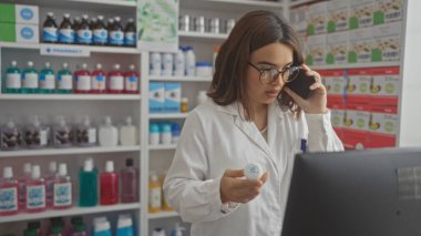 Genç ve çekici bir İspanyol esmer kadın kapalı bir eczanede laboratuvar önlüğü giyerek çalışıyor, telefonda konuşurken ilaç şişesini tutuyor ve bilgisayar ekranına bakıyor..