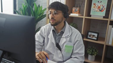 Handsome arab male doctor in headset working at clinic office with computer, paperwork, and indoor plants. clipart