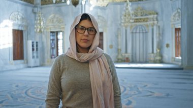 Türbanlı genç bir kadın, çarpıcı bir İstanbul camisinin içinde karmaşık bir İslami mimari ile düşünceli bir şekilde duruyor..