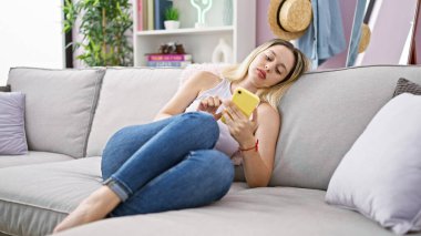 Evdeki kanepede oturan akıllı telefon kullanan genç sarışın kadın.